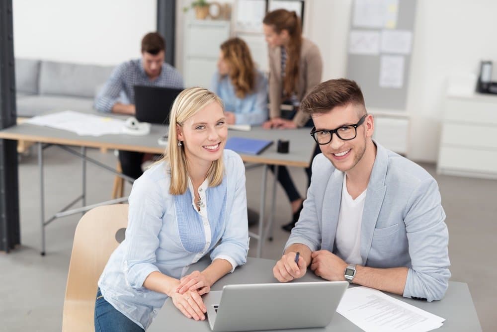 Young Office Workers Sitting at the Table with Laptop Computer Looking at the Camera with Toothy Smiles.